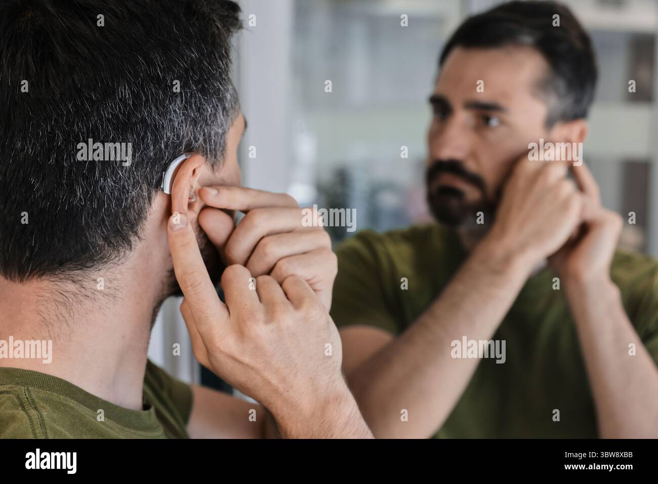 L'uomo adulto esplora il comfort e l'adattabilità di un nuovo apparecchio acustico dietro l'orecchio, regolandolo con attenzione davanti a uno specchio. Elegante apparecchio acustico Foto Stock