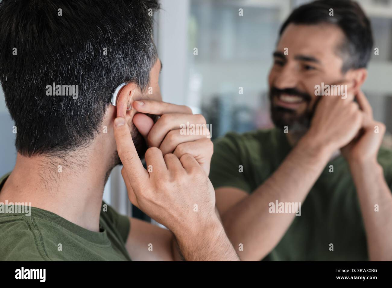Bell'uomo con apparecchio acustico BTE guarda se stessa nello specchio e prova a usare l'apparecchio acustico, primo piano. Trattamento per la perdita dell'udito Foto Stock