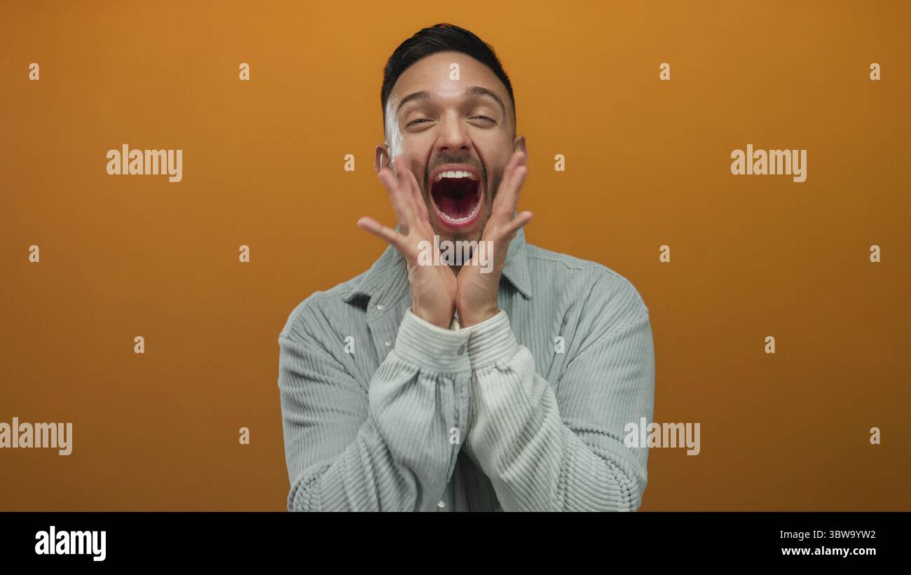 Giovane uomo che esprime gioia su un vivace sfondo arancione indossando una camicia grigia casual e sorridendo ampiamente e coinvolgendo gli spettatori con energia e unicità Foto Stock