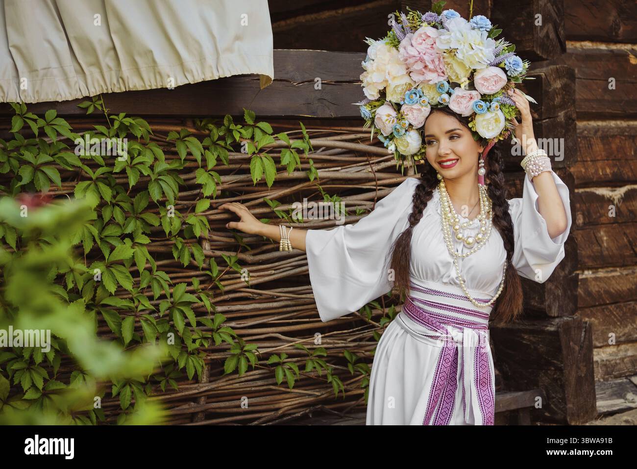 Attraente fanciulla bruna con lunghi capelli scuri, in un autentico costume nazionale ucraino bianco e una corona di fiori colorati sorride e guarda Foto Stock