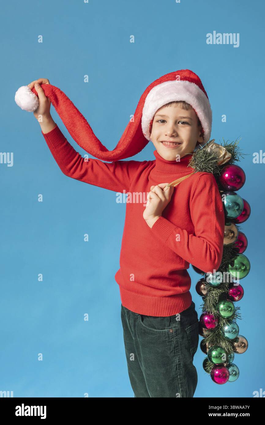 Froehliches Kind in Santa Hut halten Weihnachten dekorative Kugeln auf blauem Hintergrund, Weihnachten und Urlaub Konzept Mockup. Generazione Alpha und Foto Stock