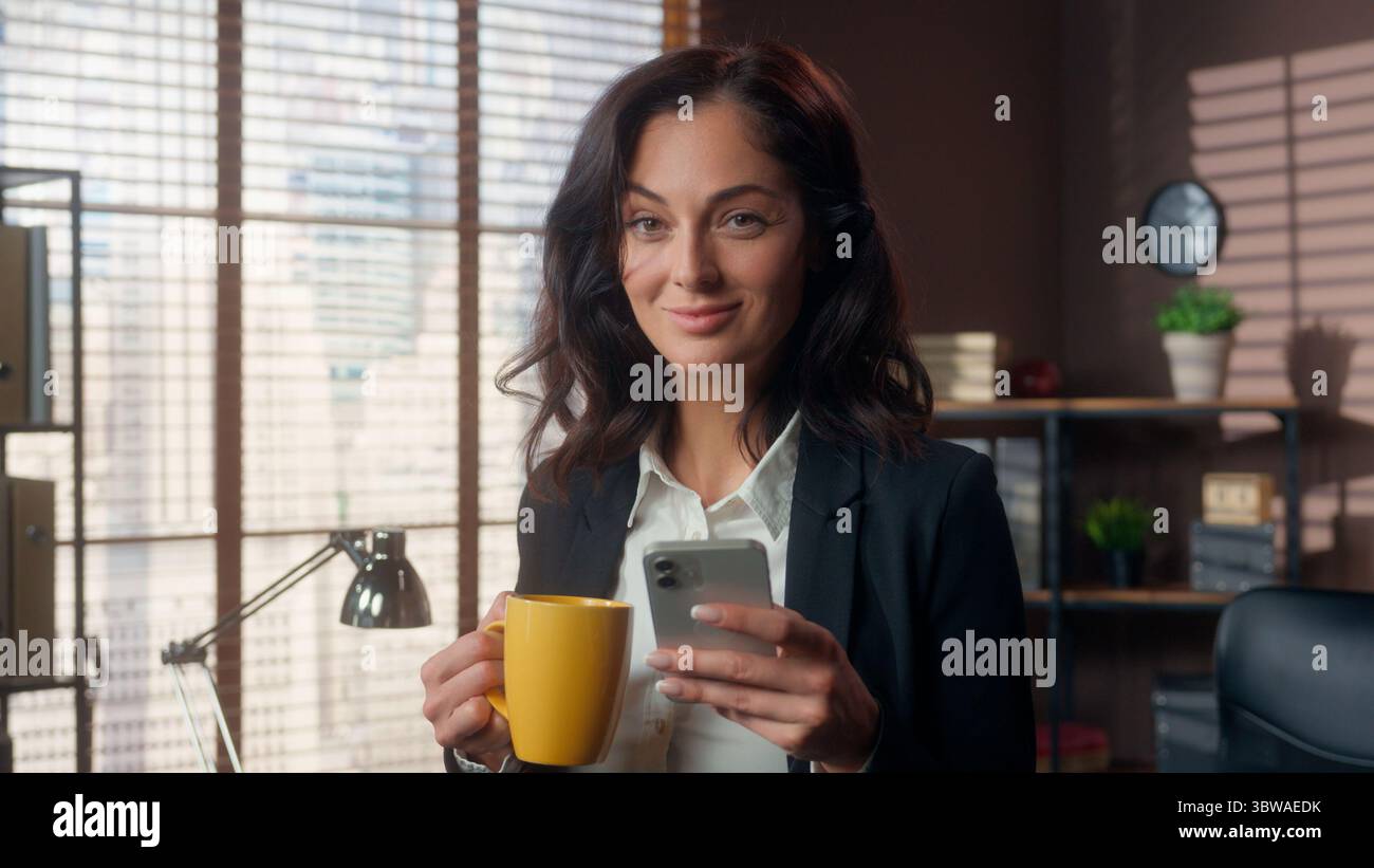 Donna caucasica che tiene una tazza di caffè utilizzando il telefono cellulare smartphone bere sorriso donna d'affari affari ufficio navigare online scorrimento Foto Stock