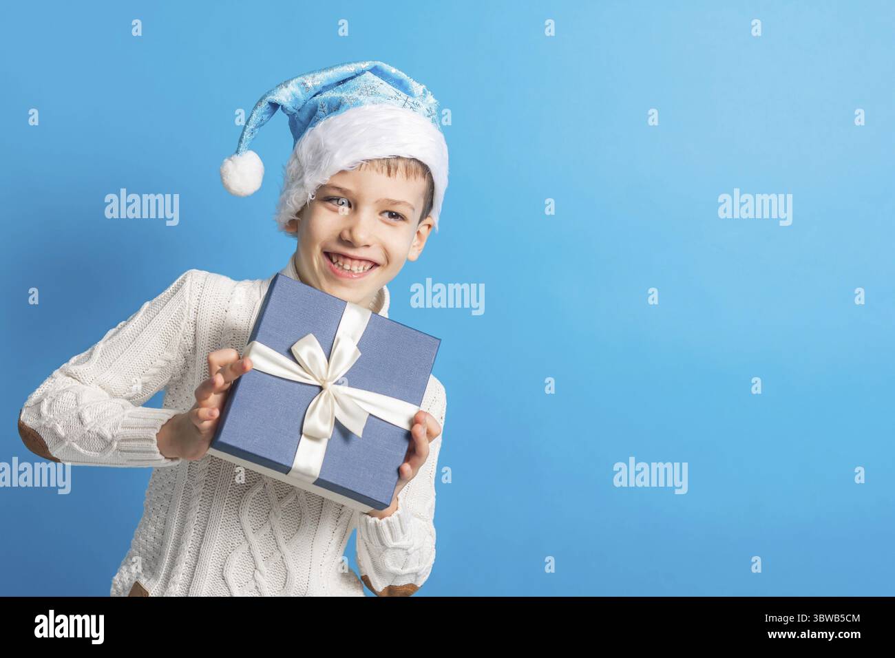 Junge haelt Geschenk. Gentile nel blauen Weihnachtshut haelt Geschenkbox. Einkaufszeit, Kopierraum und Leerraum fuer Text. Erzeugung alpha und gen alpha Foto Stock