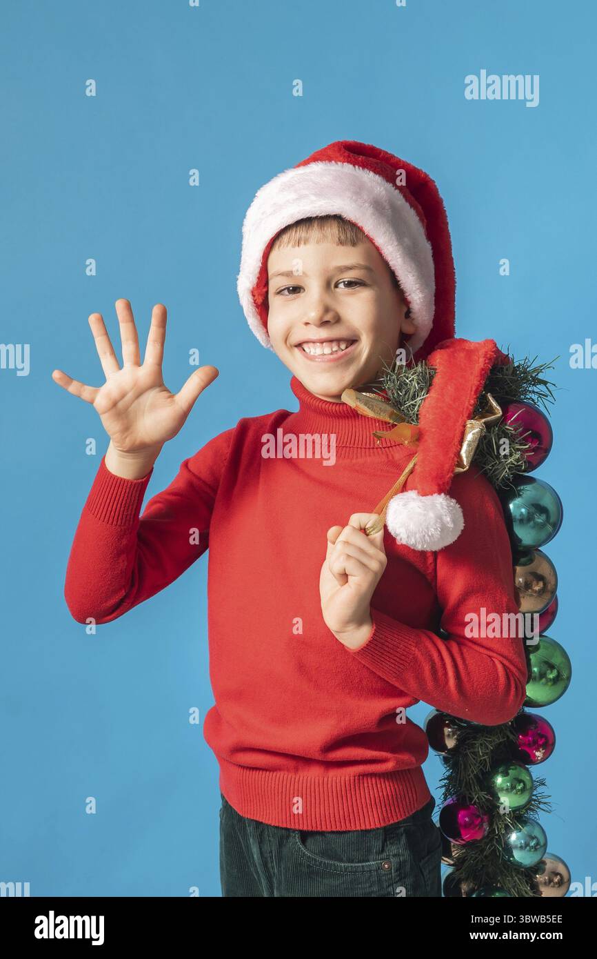 Froehliches Kind in Santa Hut halten Weihnachten dekorative Kugeln auf blauem Hintergrund, Weihnachten und Urlaub Konzept Mockup. Generazione Alpha und Foto Stock