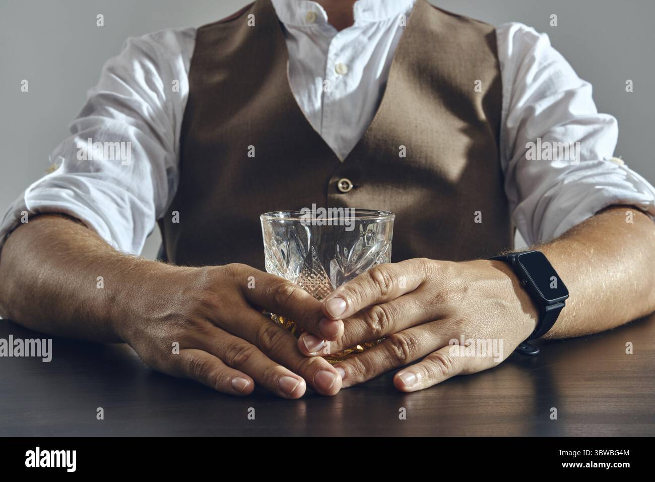 Die Haende eines Mannes in einer klassischen braunen Weste und einem weissen Hemd sitzen am Tisch und geniessen einen Whiskey. Grauer Hintergrund, Nah Foto Stock