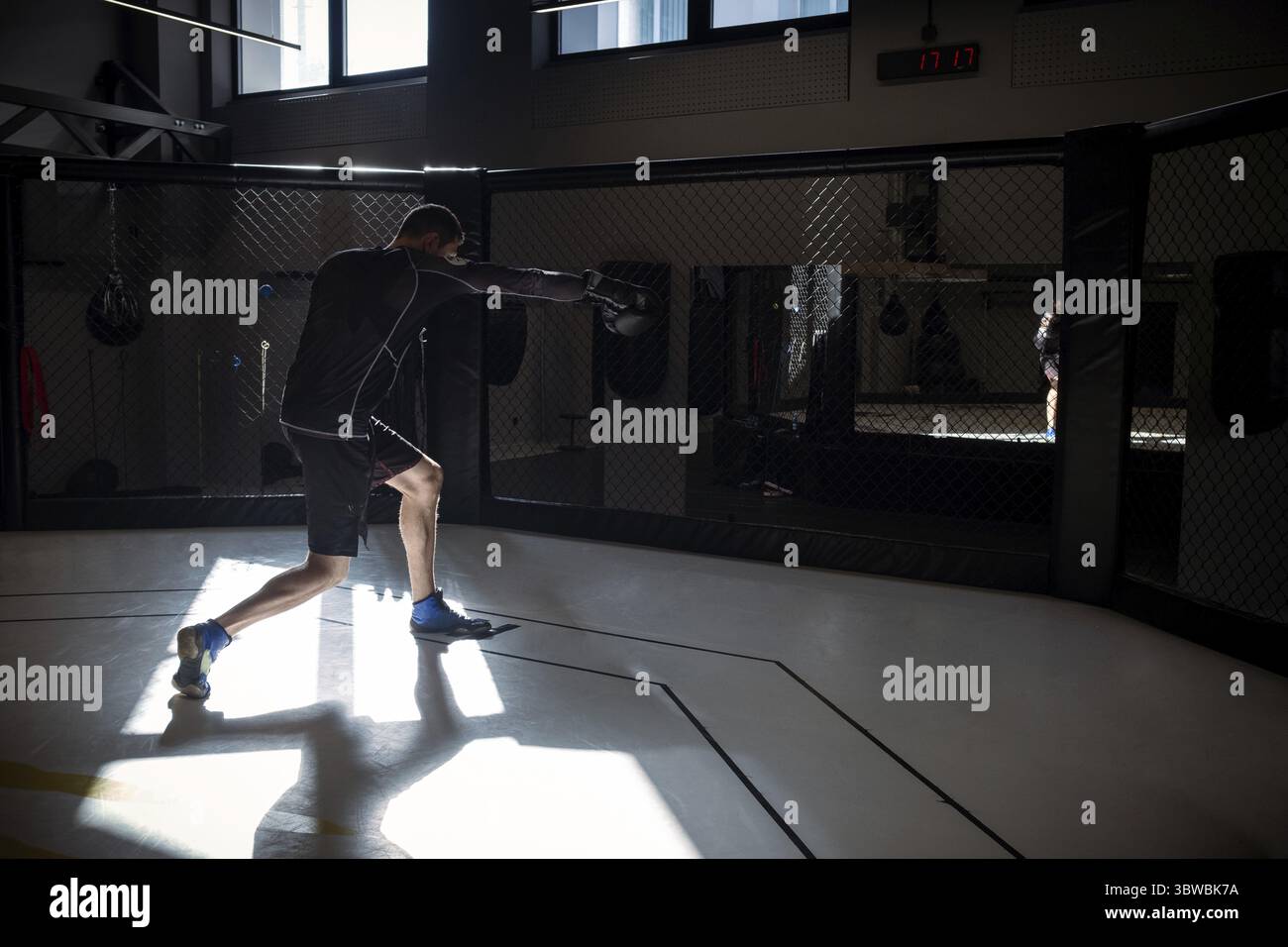 Atleta maschile dedicato impegnato in una sessione di shadowboxing, simulando mosse di combattimento contro avversari immaginari in una gabbia ottagonale di pugilato in moderni setti da palestra Foto Stock