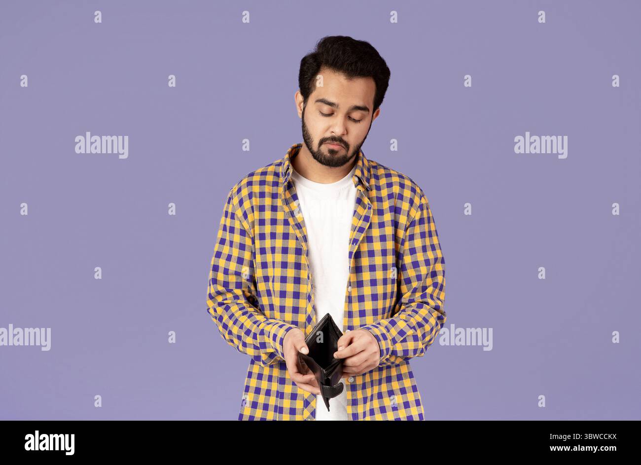 Crisi economica. Insoddisfatto ragazzo indiano che guarda nel portafoglio vuoto su sfondo viola Foto Stock