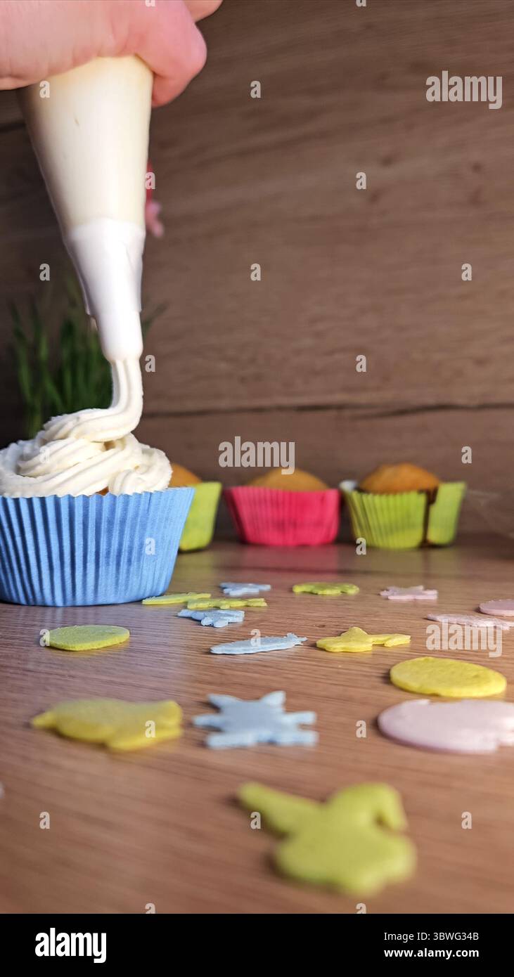 Versare a mano la panna su un cupcake in una cassa blu con custodie colorate sullo sfondo. Spruzzi decorativi sparsi su un tavolo di legno Foto Stock