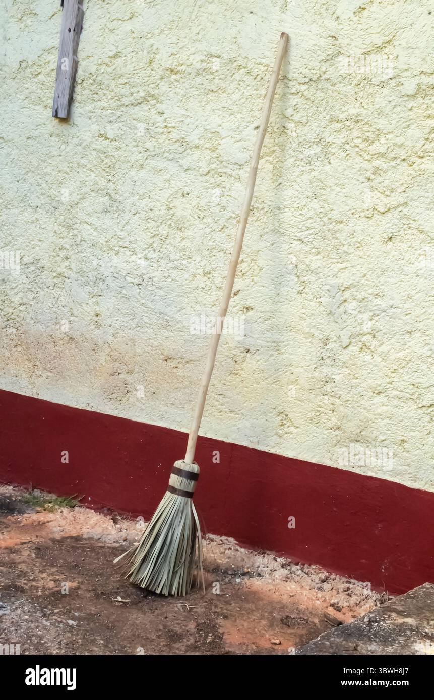 Una tradizionale scopa di paglia fatta a mano appoggiata su una rustica parete in stucco giallo con una base dipinta di rosso. Foto Stock