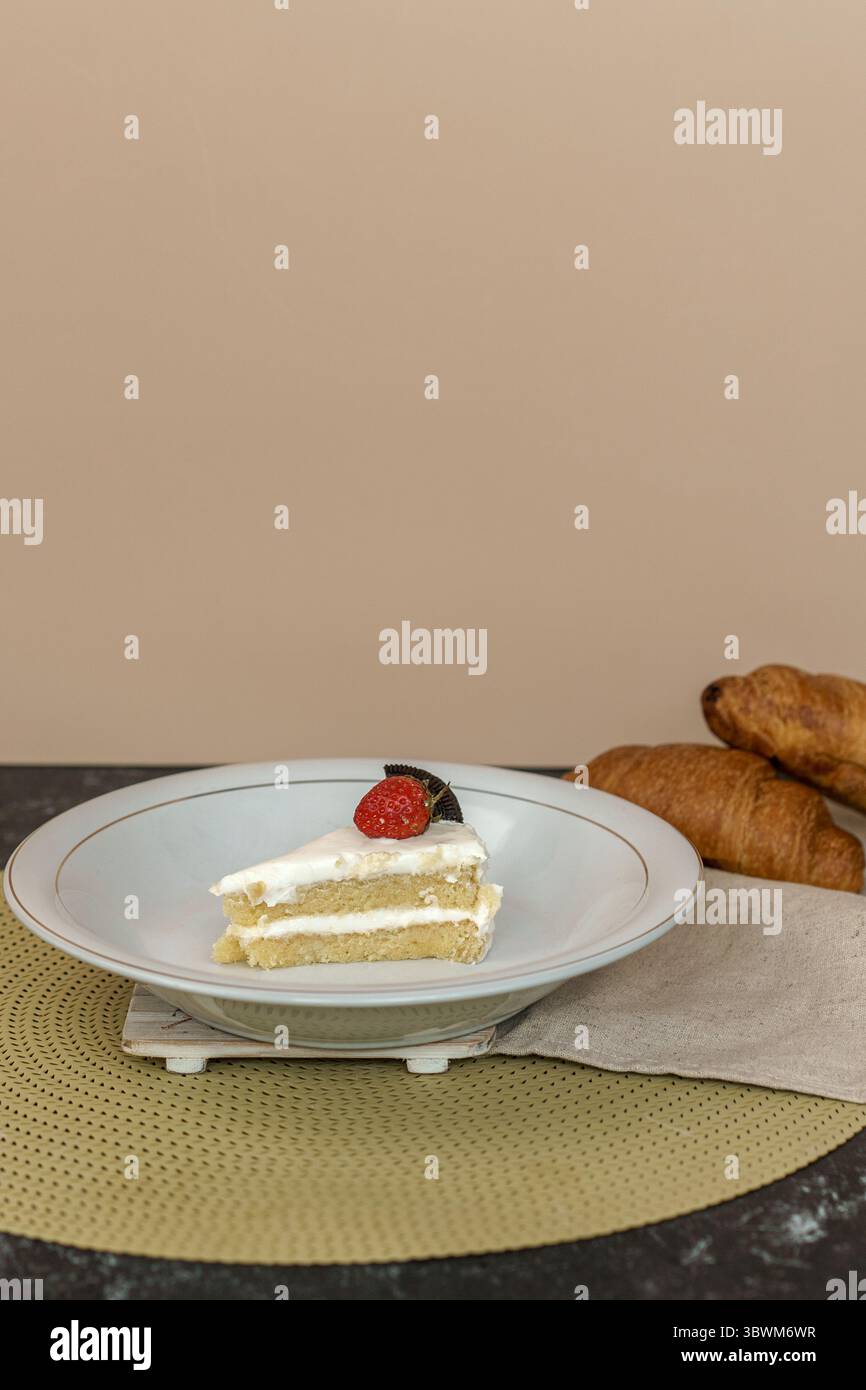 Una fetta di deliziosa torta a strati con glassa di panna bianca, condita con una fragola fresca e un biscotto. Un dessert perfetto con croissant sul retro. Foto Stock