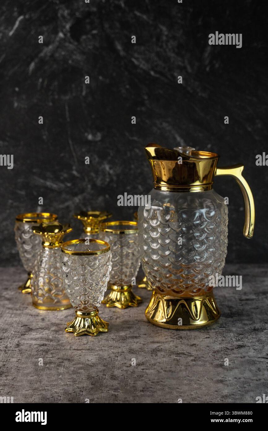 Caraffa e calice in vetro vintage. Stoviglie ornate e testurizzate con finiture dorate per una cena di lusso su uno sfondo scuro e testurizzato. Foto Stock