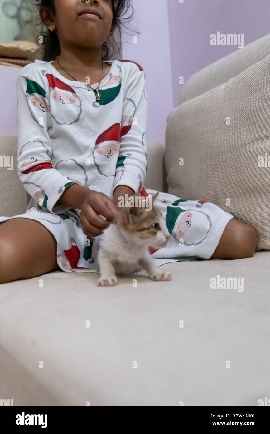 Una ragazza in pigiama festoso interagisce con gioia con un piccolo gattino su un divano, creando un'atmosfera calda e giocosa. Foto Stock