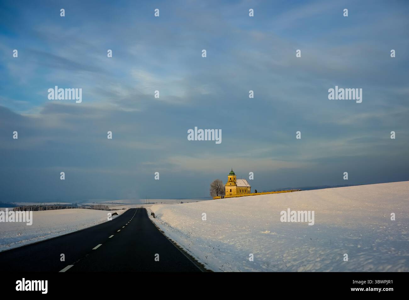 La strada di campagna innevata conduce oltre una piccola chiesa illuminata dalla calda luce solare invernale sotto un cielo spettacolare. Foto Stock