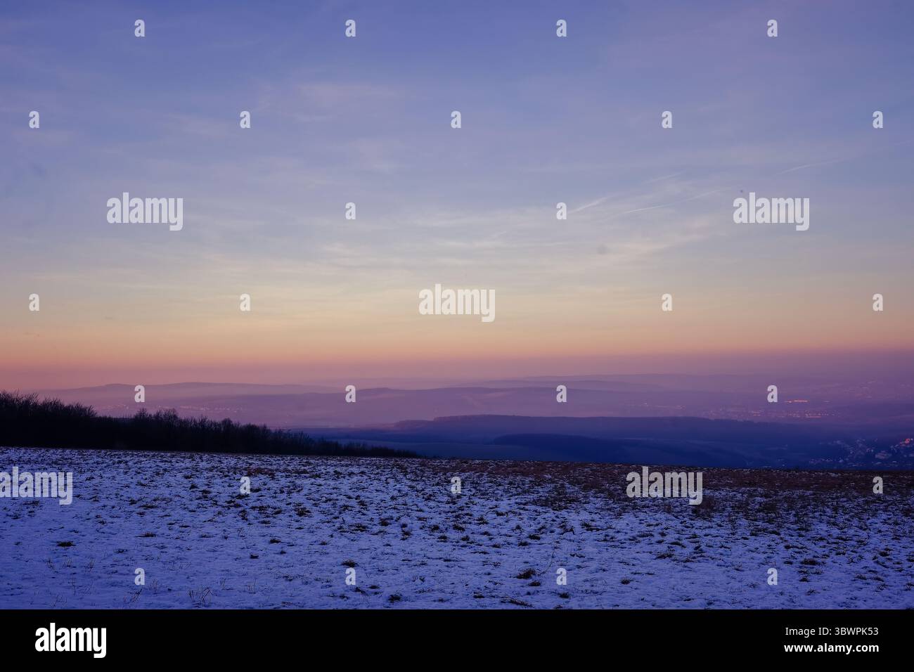 Paesaggio invernale con campi innevati e colline stratificate al tramonto, caratterizzato da un cielo sfumato in tenui colori pastello dall'arancione al viola. Calma e atmosfera Foto Stock