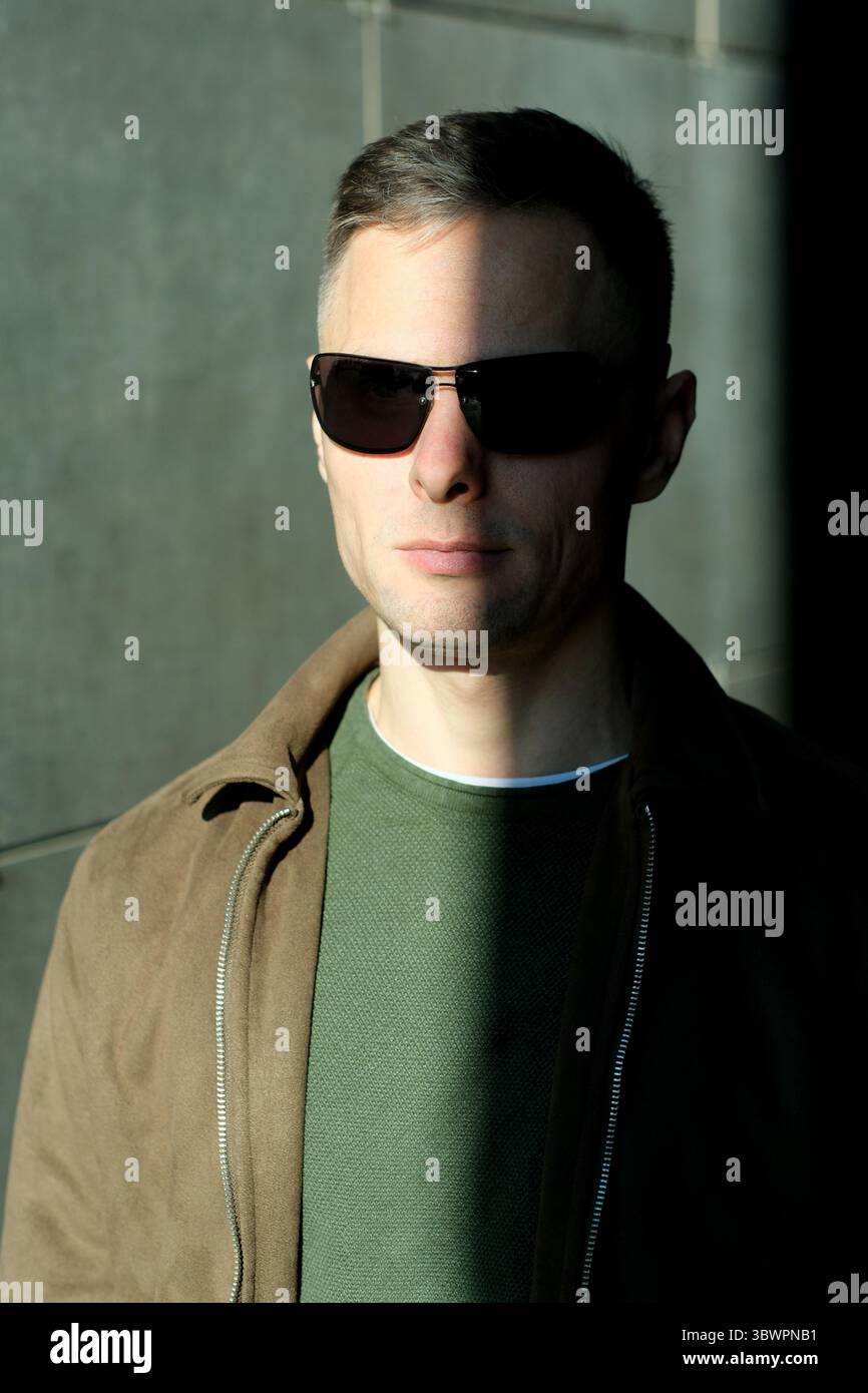 Un uomo sicuro di sé che indossa gli occhiali da sole sta alla luce del sole, gettando ombre sul viso. Foto Stock
