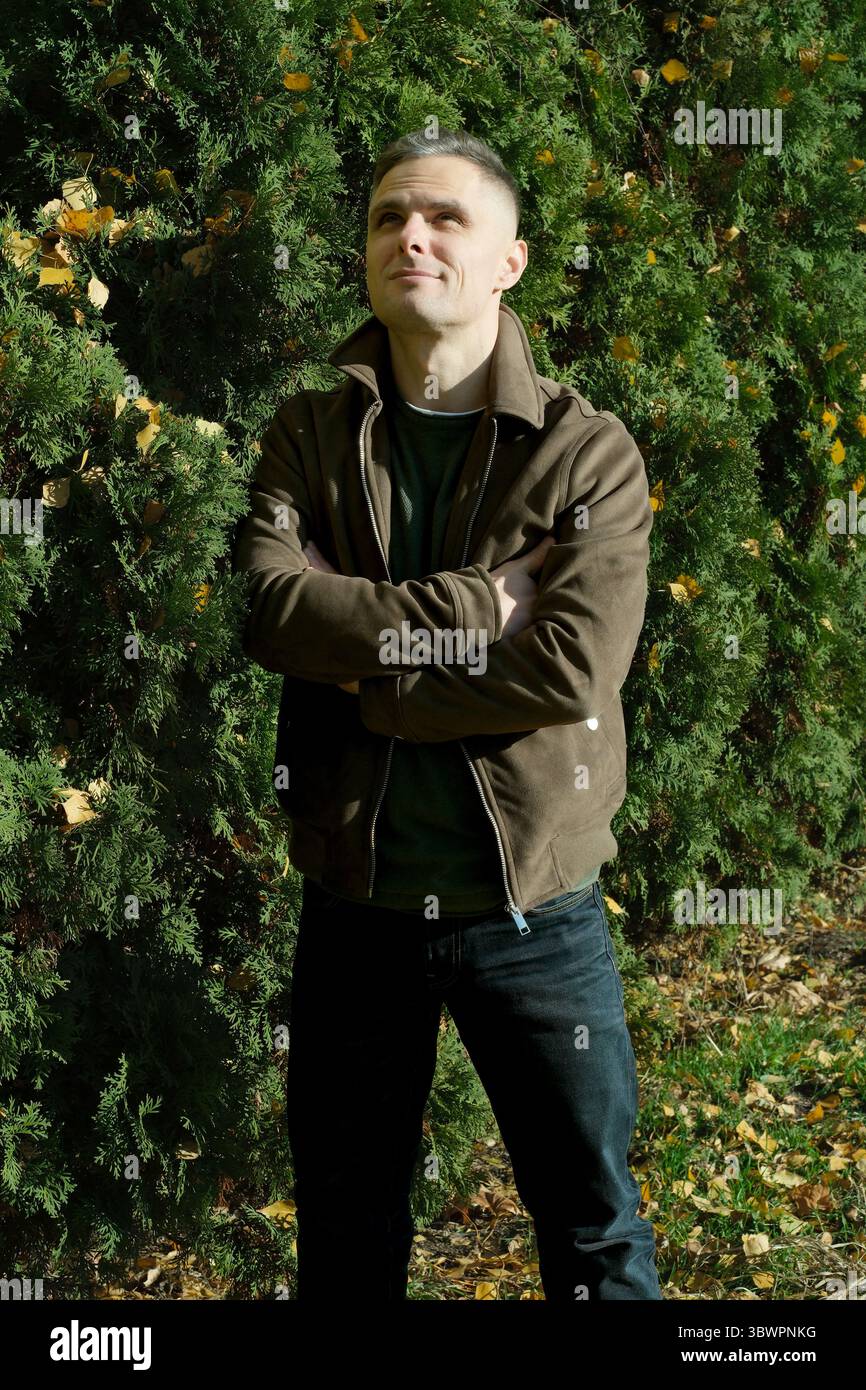 Un uomo allegro sta con le braccia incrociate, immergendosi nell'atmosfera autunnale del giardino. Foto Stock