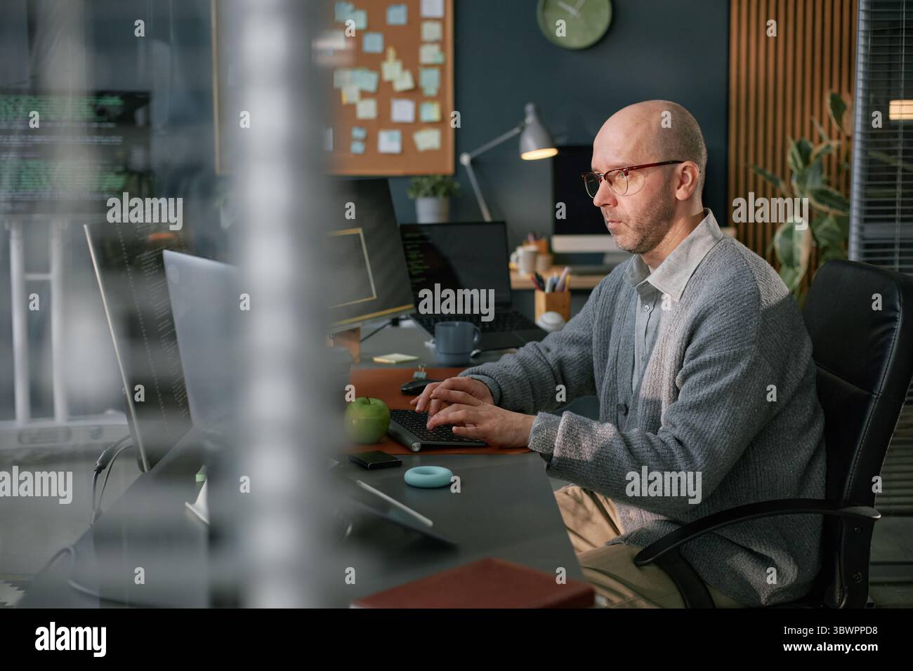 Uomo caucasico di mezza età che lavora al computer in un ufficio moderno, digita la tastiera e si concentra sullo schermo, circondato da dispositivi tecnologici e forniture per ufficio Foto Stock