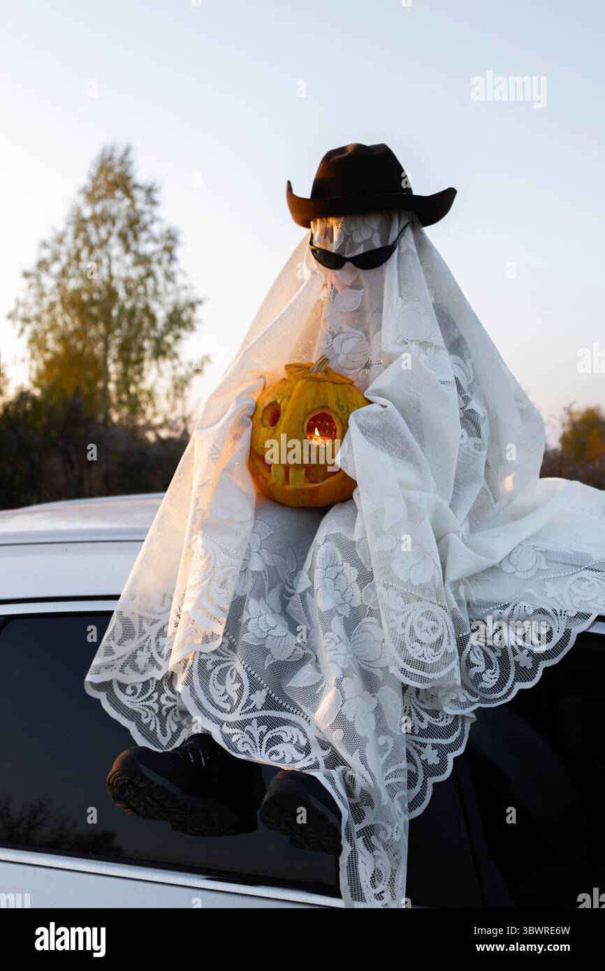 il bambino con coperta bianca, cappello, occhiali neri con una zucca arancione scolpita per le vacanze siede sul tetto di un'auto bianca. Persone in costumi fantasma per Halloween Foto Stock