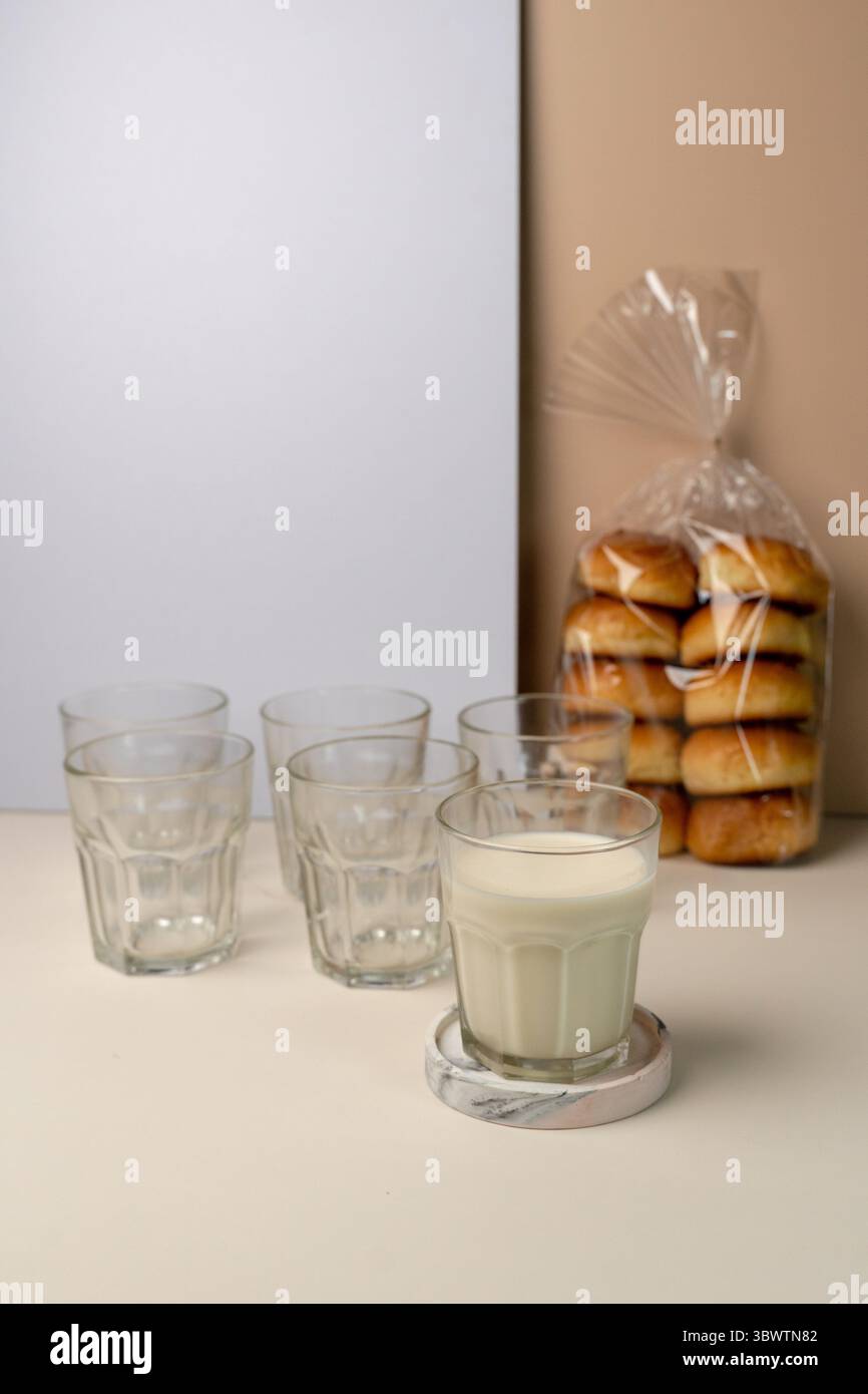 Bicchiere di latte fresco su un coaster in marmo, pronto per una sana colazione. Una confezione di panini morbidi e bicchieri vuoti sullo sfondo. Foto Stock