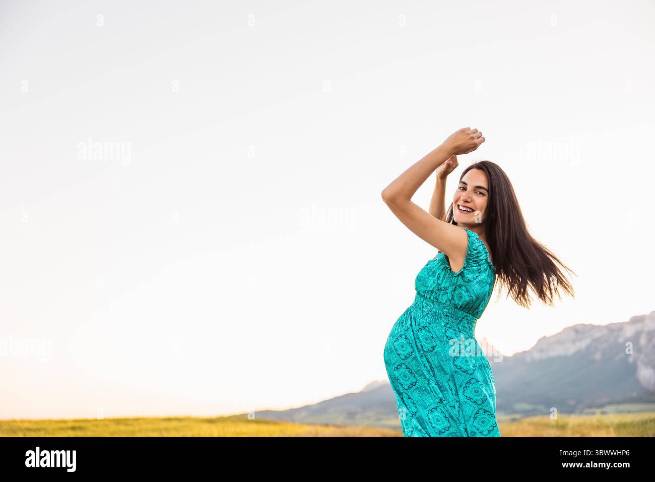 Una donna incinta in un abito turchese sorride brillantemente con le braccia alzate, abbracciando il momento in un ambiente panoramico all'aperto con campi dorati Foto Stock