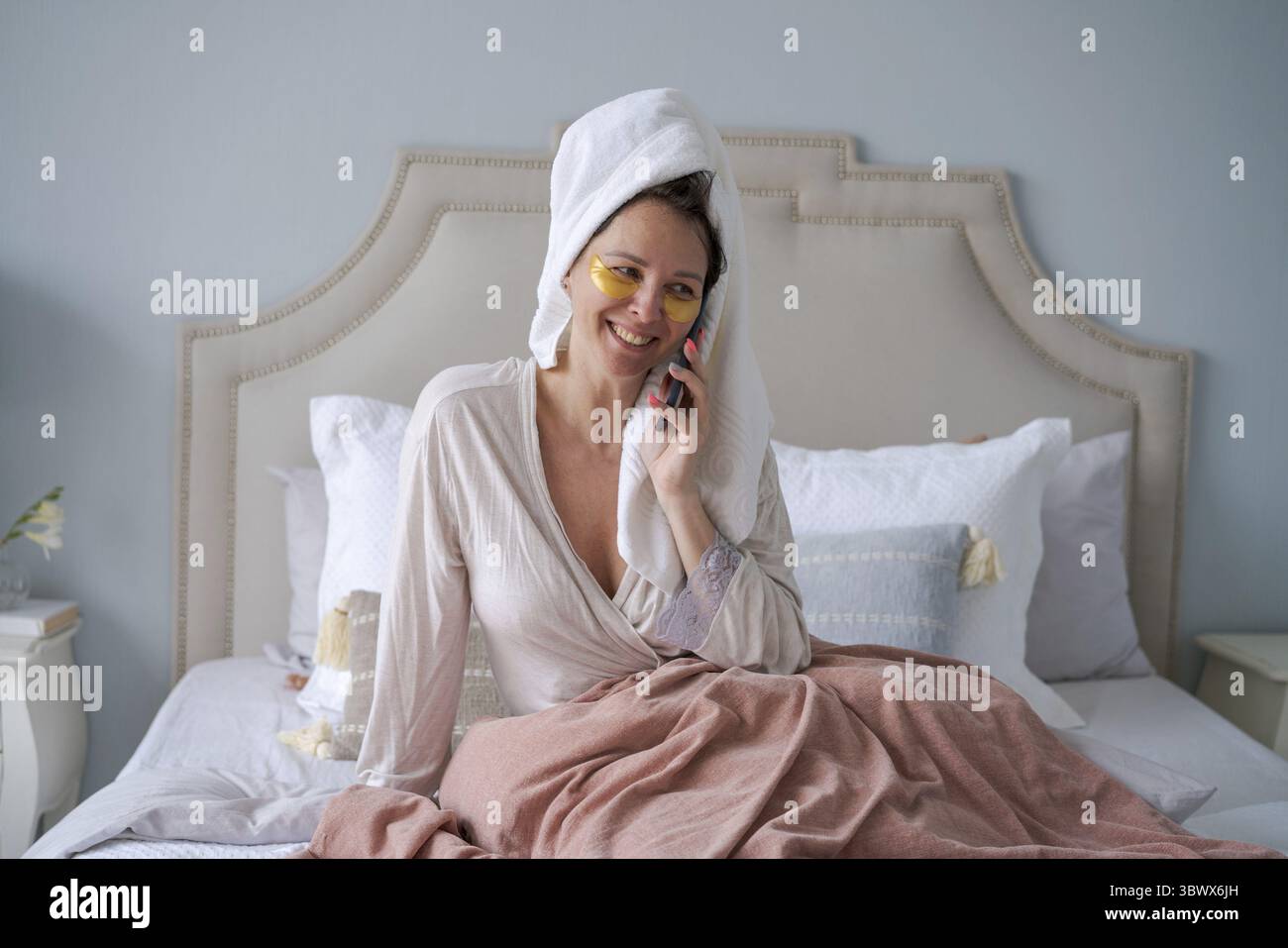 Bella donna con la benda e con il telefono cellulare comunica tramite connessione cellulare in un accappatoio e un asciugamano sulla testa, sdraiata a letto. Beaut Foto Stock