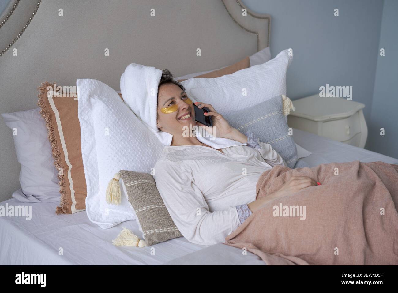 Bella donna con la benda e con il telefono cellulare comunica tramite connessione cellulare in un accappatoio e un asciugamano sulla testa, sdraiata a letto. Beaut Foto Stock