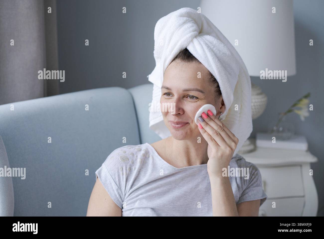 Donna sorridente con un asciugamano sulla testa e in accappatoio che regge un tampone di cotone che pulisce la pelle del viso con il detergente, happy lady rimuove il look da trucco e Foto Stock