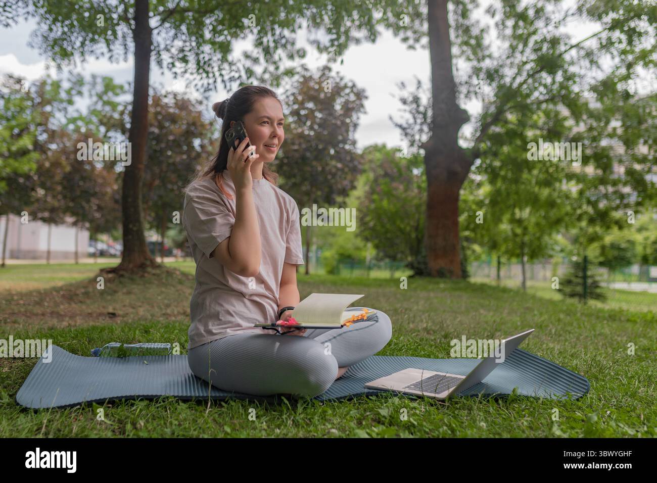 Donna sorridente che tiene in mano lo smartphone che parla all'aperto seduta su un tappetino yoga a gambe incrociate notebook aperto argento, parco verde, sfondo verde. Foto Stock