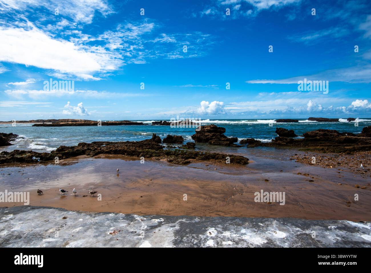 Le rocce intorno alla città di Essaouira proteggono la città marocchina dall'Oceano Atlantico tra cieli blu e nuvole bianche Foto Stock