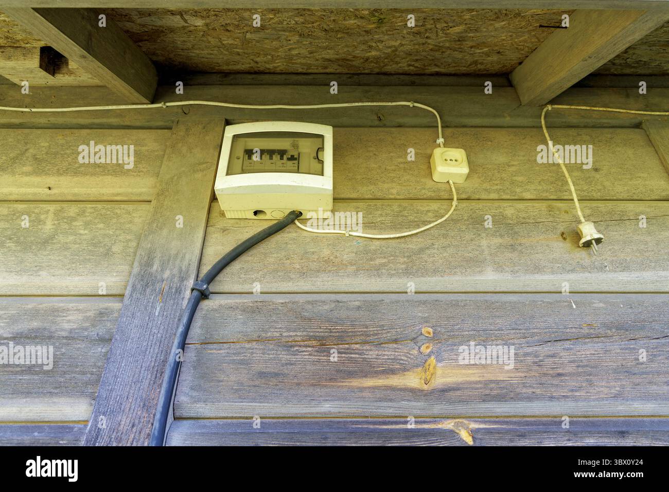 Una scatola elettrica con fili e una spina montata su una parete di legno intemprata sotto un semplice soffitto di legno, che mostra un'utilità rustica. Foto Stock