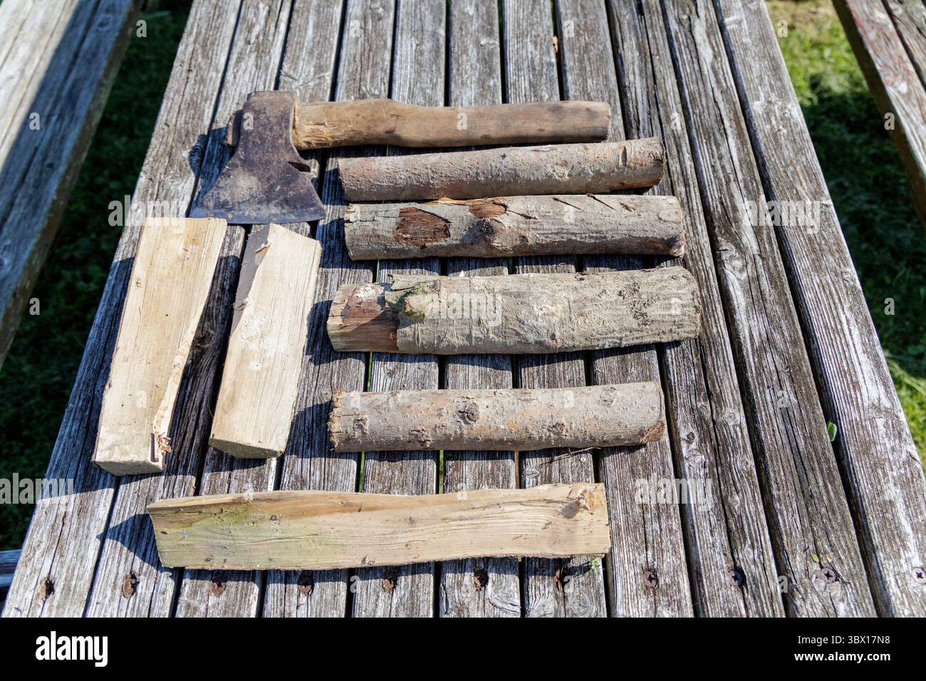 Una vecchia ascia e vari pezzi di legna da ardere sono disposti su una tavola rustica di legno, suggerendo la preparazione per un fuoco o lavori all'aperto. Foto Stock