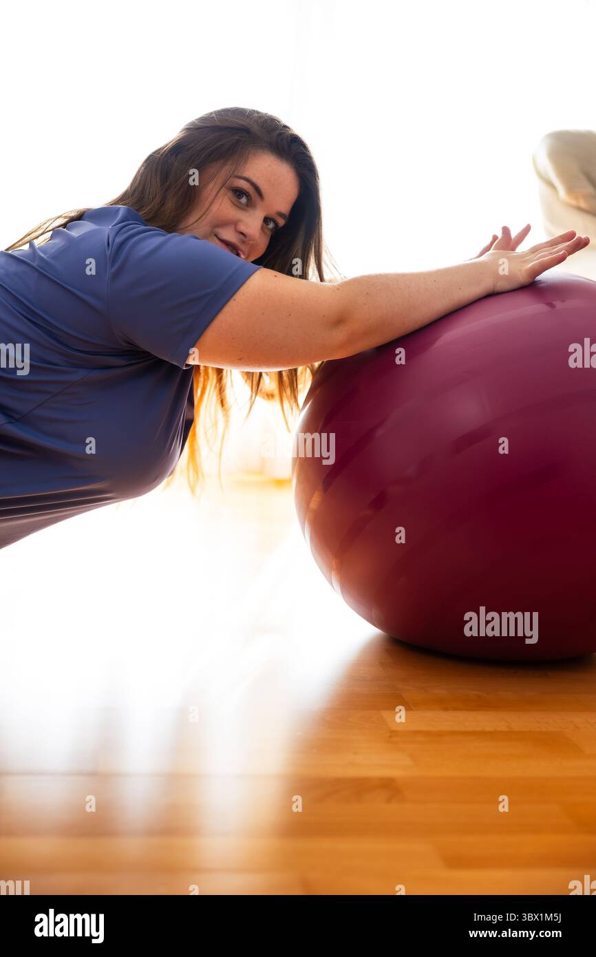 Primo piano di una donna incinta con abiti da ginnastica inginocchiati sul pavimento e appoggiati a una palla da fitness, sorridendo delicatamente in un ambiente domestico con luce soffusa. Foto Stock