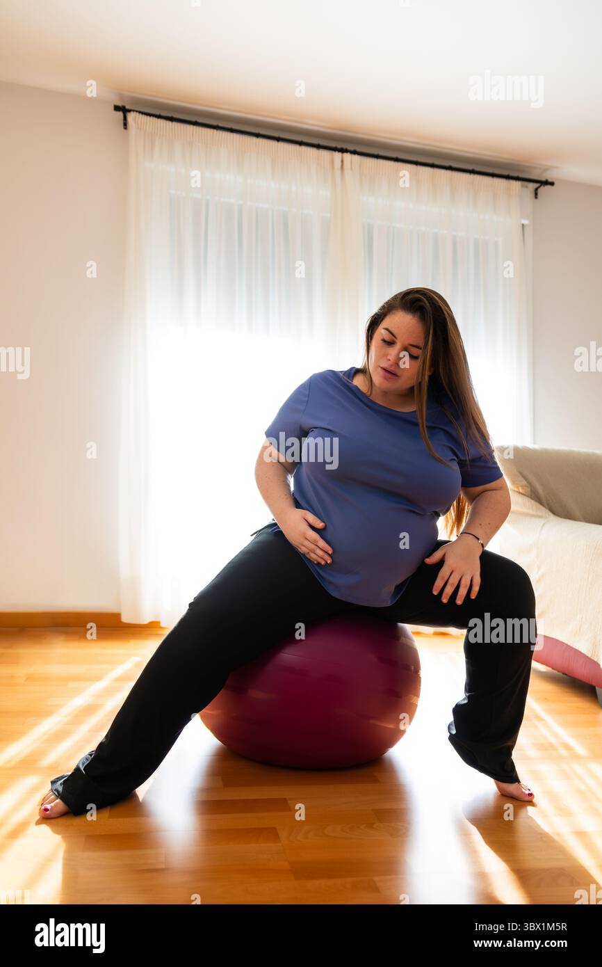 Donna incinta che indossa abbigliamento sportivo seduto su una palla da ginnastica, che si allunga le gambe e tiene la pancia in un ambiente domestico illuminato dal sole con una lastra di legno Foto Stock