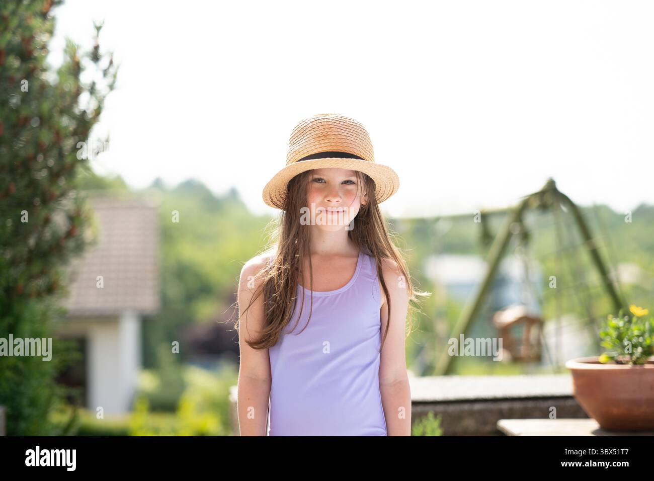 Sorridente ragazza con cappello di paglia e costume da bagno su un balcone estivo. Concetto: Felicità dell'infanzia, atmosfera di vacanza, gioia del sole, momenti all'aperto spensierati. Foto Stock