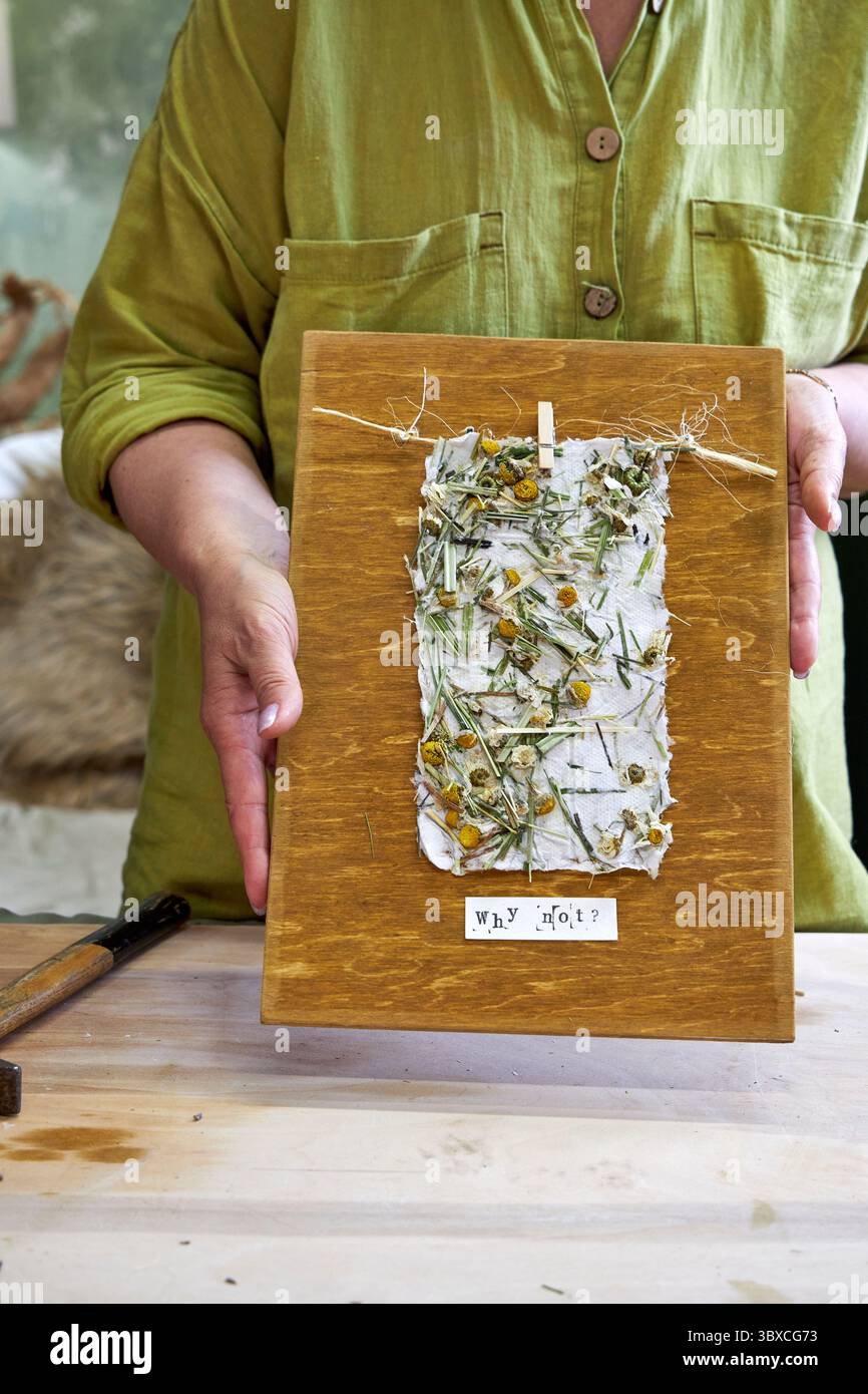 Donna che tiene in mano un pannello da parete finito in carta floreale fatta a mano su legno. Artigianato ecologico con materiali naturali e messaggio positivo: "Perché no? Foto Stock Donna che tiene in mano un pannello da parete finito in carta floreale fatta a mano su legno. Artigianato ecologico con materiali naturali e messaggio positivo: "Perché no? Foto Stock