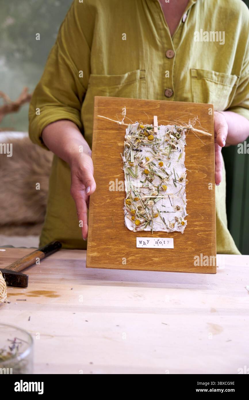 Donna che tiene in mano un pannello da parete finito in carta floreale fatta a mano su legno. Artigianato ecologico con materiali naturali e messaggio positivo: "Perché no? Foto Stock Donna che tiene in mano un pannello da parete finito in carta floreale fatta a mano su legno. Artigianato ecologico con materiali naturali e messaggio positivo: "Perché no? Foto Stock