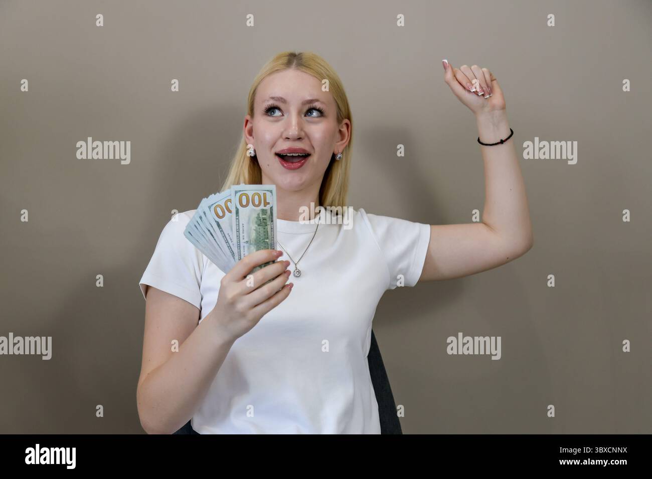 Donna bionda con una t-shirt bianca che regge gioiosamente una pila di banconote da cento dollari e che si alza il braccio per festeggiare, esprimendo il successo finanziario e.. Foto Stock