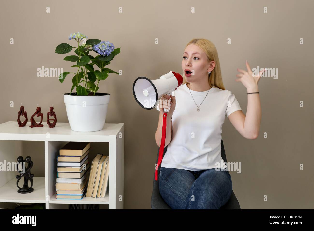 Donna bionda con t-shirt casual bianca e jeans gestiscono con passione parlando in un megafono rosso e bianco, seduti in un intimo interno con bo Foto Stock