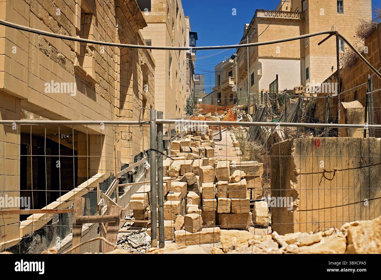 Cantiere urbano ingombrante con blocchi di pietra calcarea, recinzioni metalliche e tavole di legno che bloccano una strada stretta in una giornata di sole. Foto Stock
