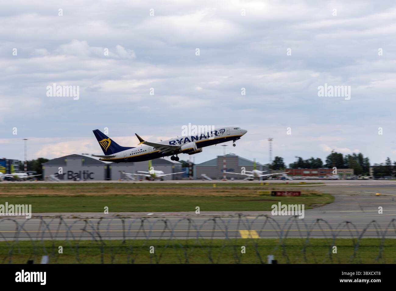 RIGA/LETTONIA – 07.06.2025: Un aeromobile Ryanair decolla dall’aeroporto internazionale di riga, con il terminal aeroportuale e la torre di controllo sullo sfondo, Foto Stock