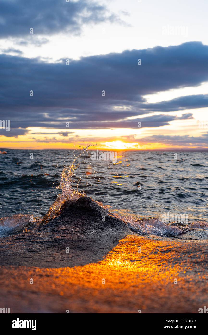 Il sole tramonta su un lago ondulato, emettendo una calda luce arancione e riflessi su una roccia in primo piano con spruzzi d'acqua. Foto Stock