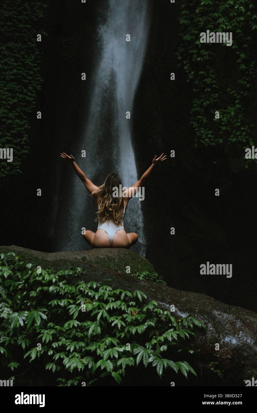 Giovane donna che medita di fronte alla maestosa cascata nella lussureggiante giungla Foto Stock