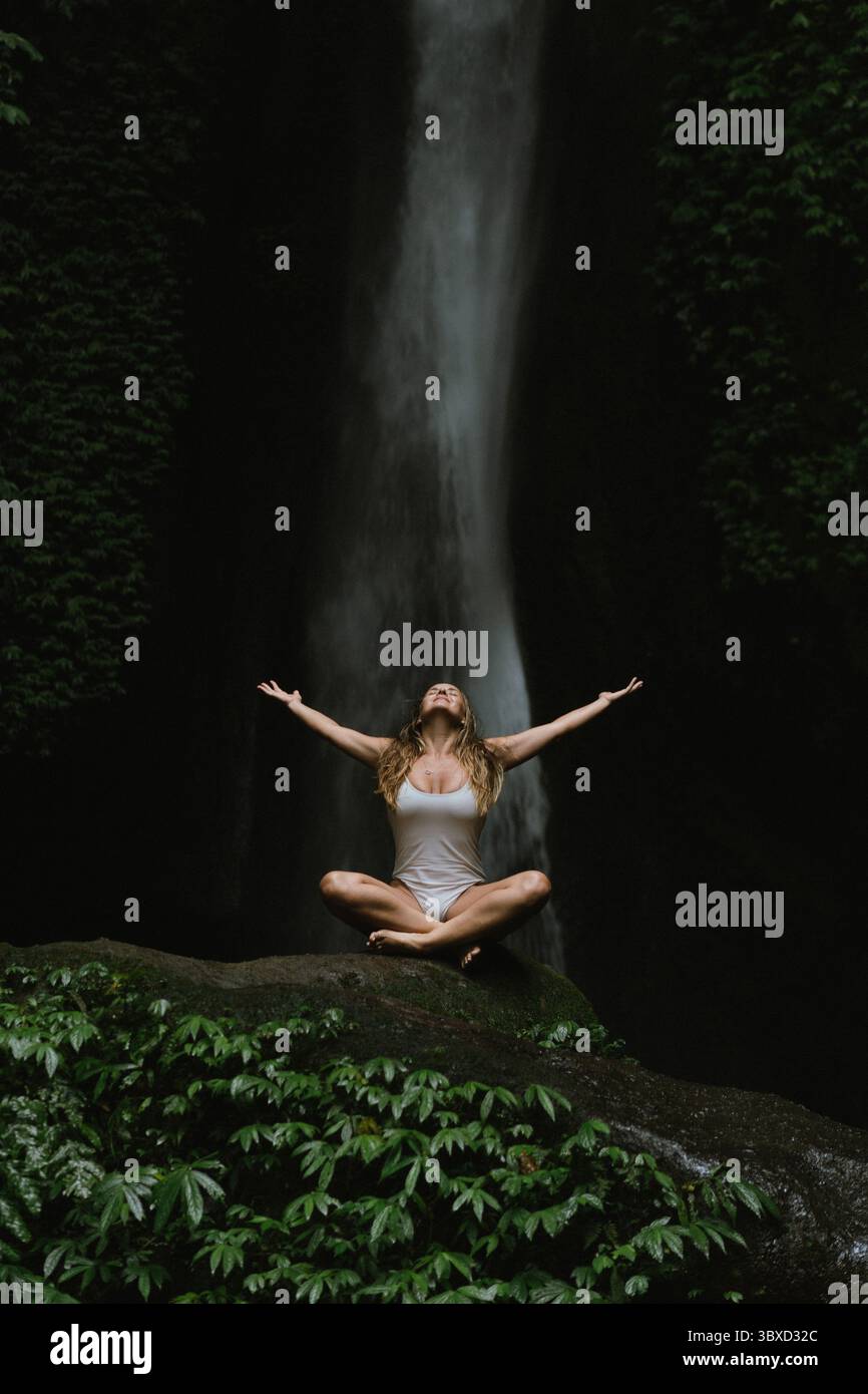 Giovane donna che medita di fronte alla maestosa cascata nella lussureggiante giungla Foto Stock
