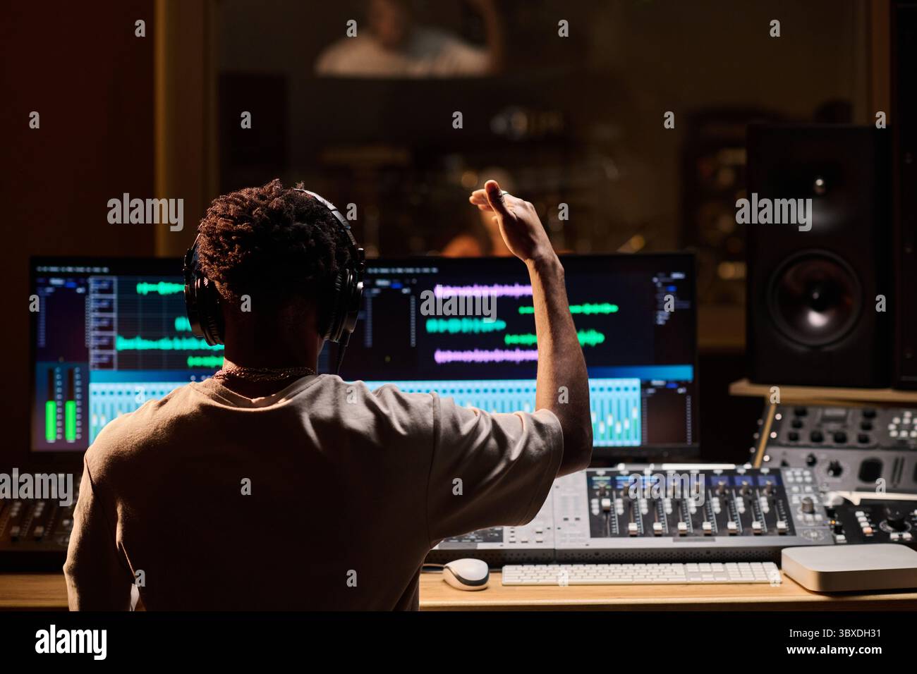 Giovane uomo adulto di colore che indossa le cuffie che lavora alla console di missaggio audio in uno studio di registrazione professionale, alzando la mano durante l'editing di brani musicali sullo schermo del computer Foto Stock