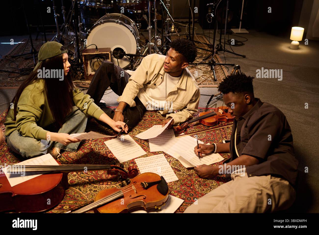 Gruppo multietnico di musicisti che collaborano alla composizione musicale, seduti a terra con violini e spartiti, discutendo idee e scrivendo note in studio di prova Foto Stock