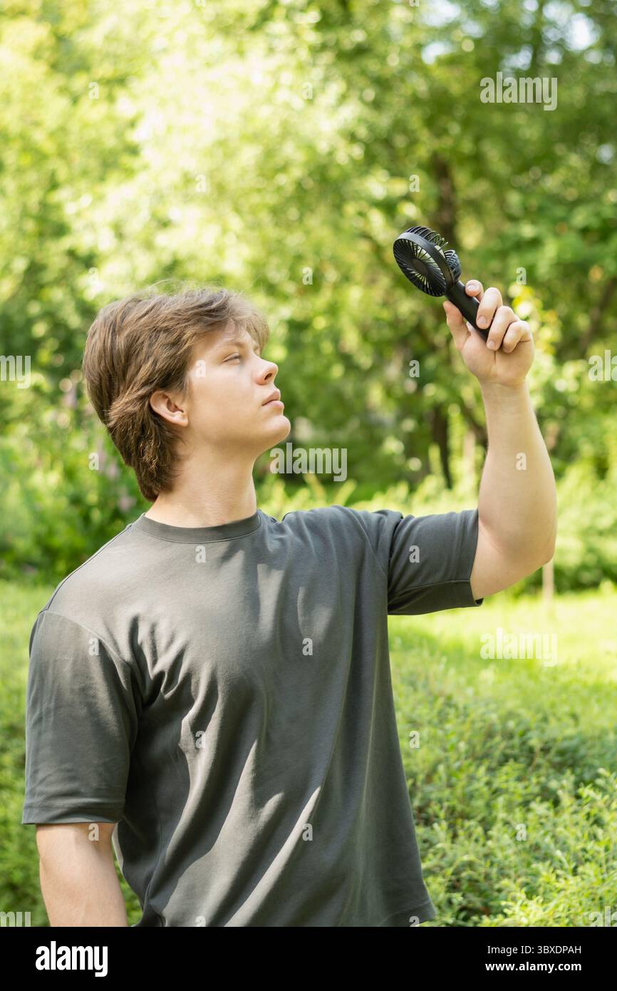 Bel giovane surriscaldato con ventilatore elettrico a mano che soffre di calore al parco. Estate calda. Ventola portatile. Sconfiggi il calore. Foto Stock
