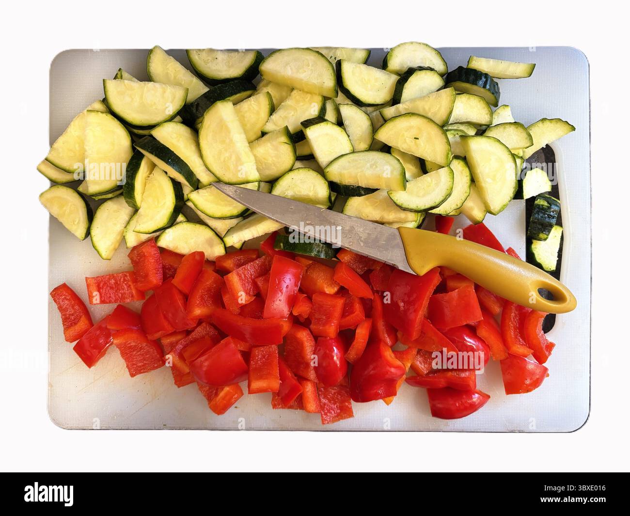 Le zucchine tagliate a fette in mezze lune e i peperoni rossi tritati riposano su un tagliere bianco con un coltello da cucina. Foto Stock