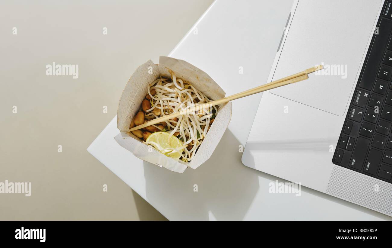 Una ciotola di tagliatelle di cartone con verdure e lime si trova accanto a un computer portatile su un tavolo bianco. L'ambiente suggerisce il multitasking, combinando la ristorazione e il Foto Stock