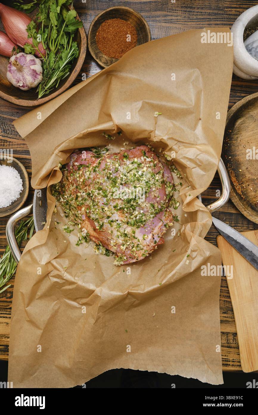 Coscia di tacchino condita in una teglia rivestita di carta pergamena, circondata da erbe fresche, spezie e altri ingredienti. Questa cucina accogliente irradia calore Foto Stock