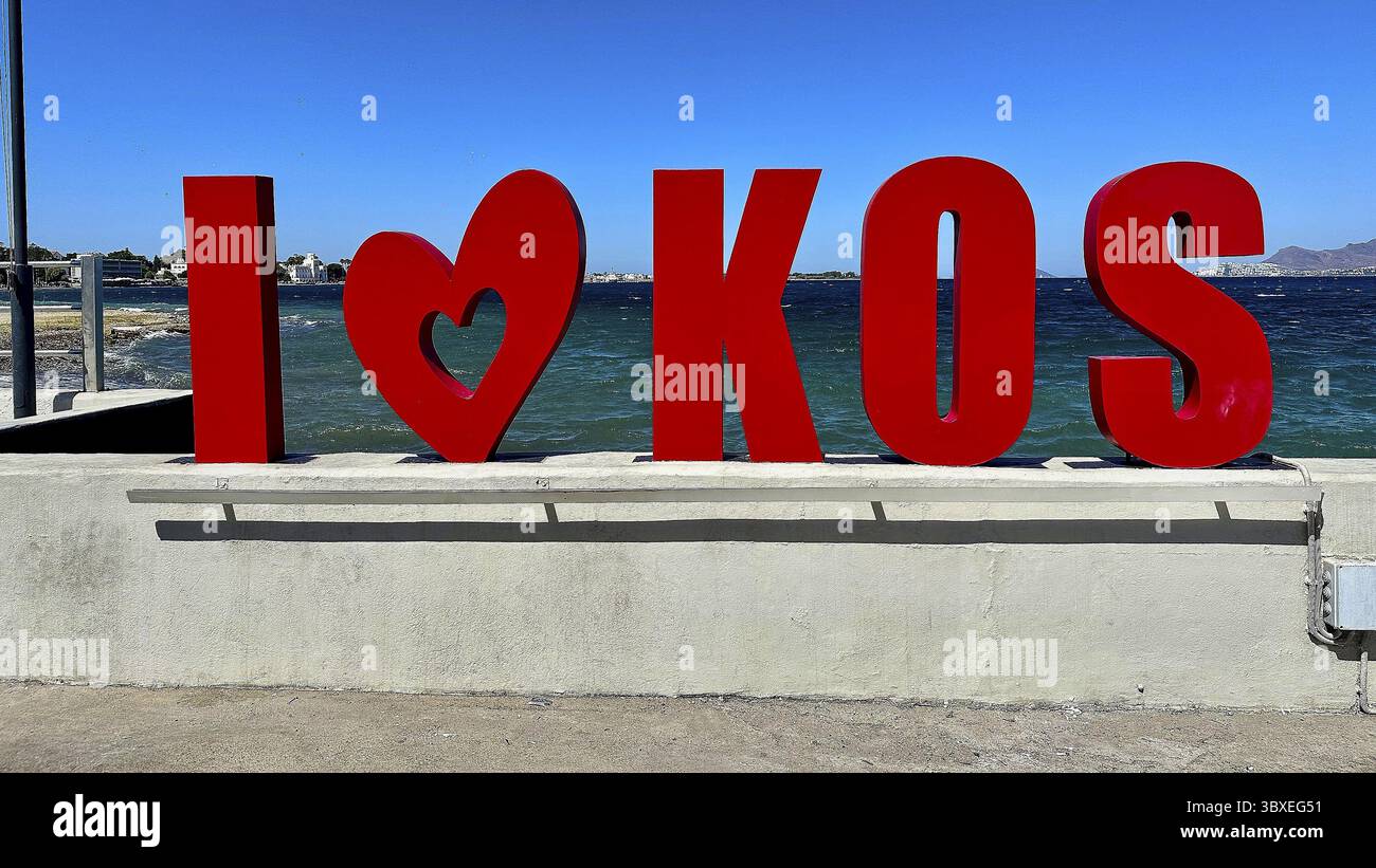 Scritta rossa "Iâ ¤ai¸ KOS" di fronte a un mare blu, moderno stile tipografico sotto un cielo estivo limpido, città di Kos, Kos, Dodecaneso, isole greche, Grecia Foto Stock